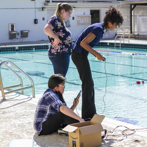 Photo of three students standing at the edge of a swimming pool, controlling an aquatic robot (offscreen) by wire.