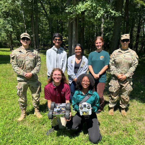 Photo of student interns holding a four-wheeled robot and a remote controller, flanked by US Army Uniformed Personnel